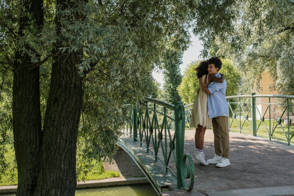 A youthful couple shares a romantic hug on a scenic park bridge under leafy trees.