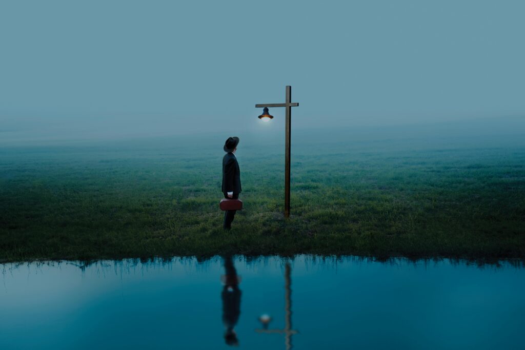 A solitary figure stands under a light post by water, reflecting solitude and hope.